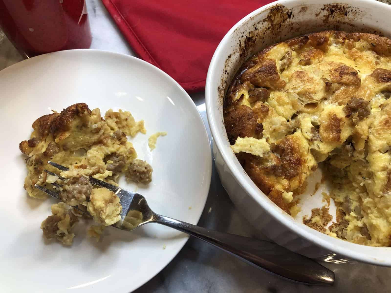 A fork resting on a white plate with a serving of egg, sausage and cheese strata and next to a white casserole dish with the egg, sausage and cheese strata with one slice missing.