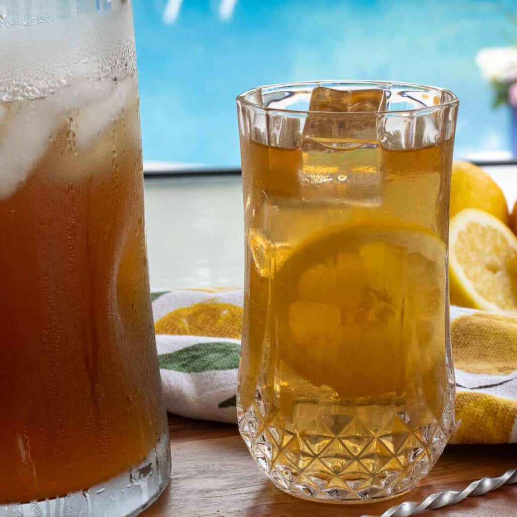A glass of Long Island Iced Tea next to a pitcher, sitting on a poolside table.