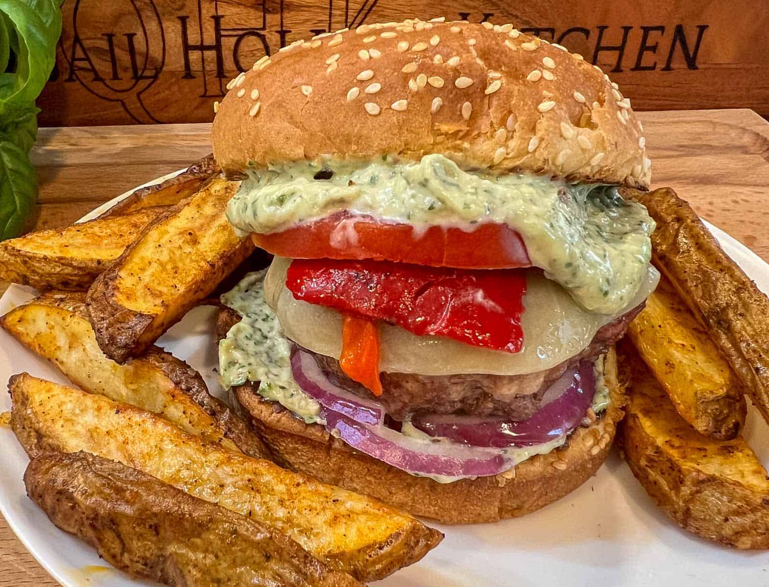 An Italian Burger on a sesame bun, topped with pesto aioli, roasted red peppers, tomato, onion and cheese, on a white plate with steak fries.