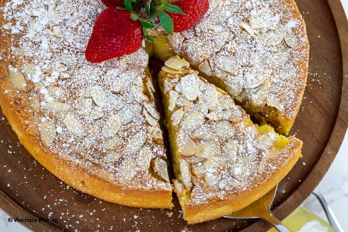 A almond cake topped with sliced almonds confectioners' sugar and strawberries with one lice being removed.