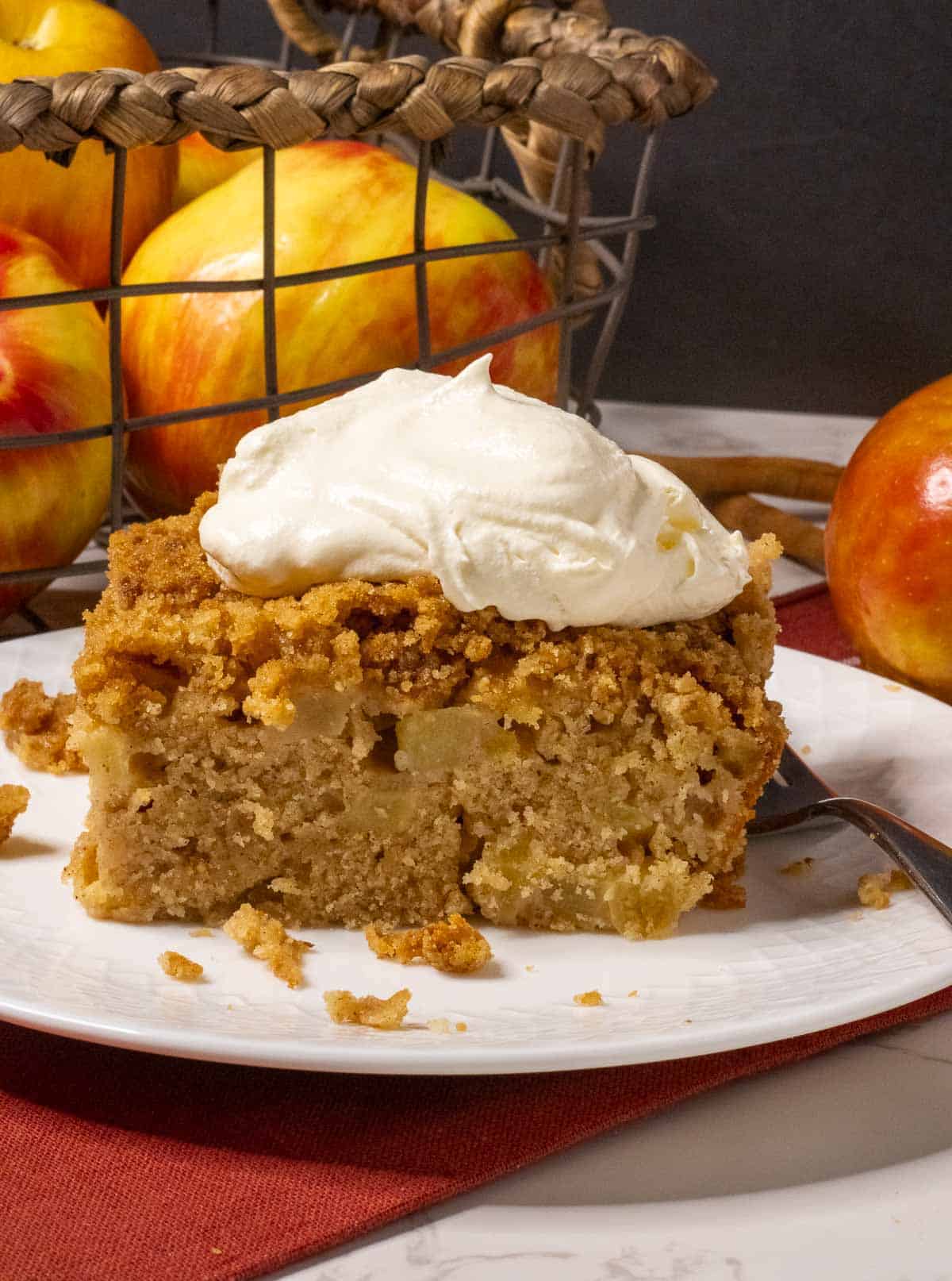 A piece of apple cake with whipped cream in front of a wire basket of apples.