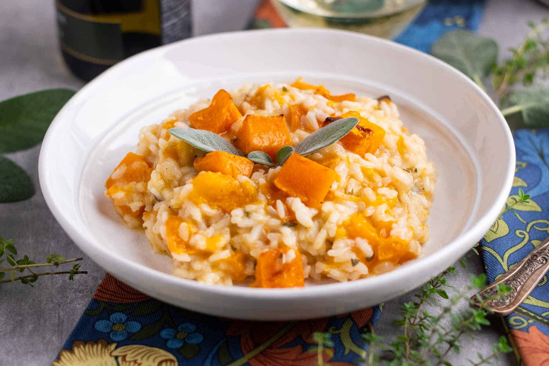 A bowl of butternut squash risotto on a table with a glass of white wine, fresh sage and thyme leaves and floral napkins.   