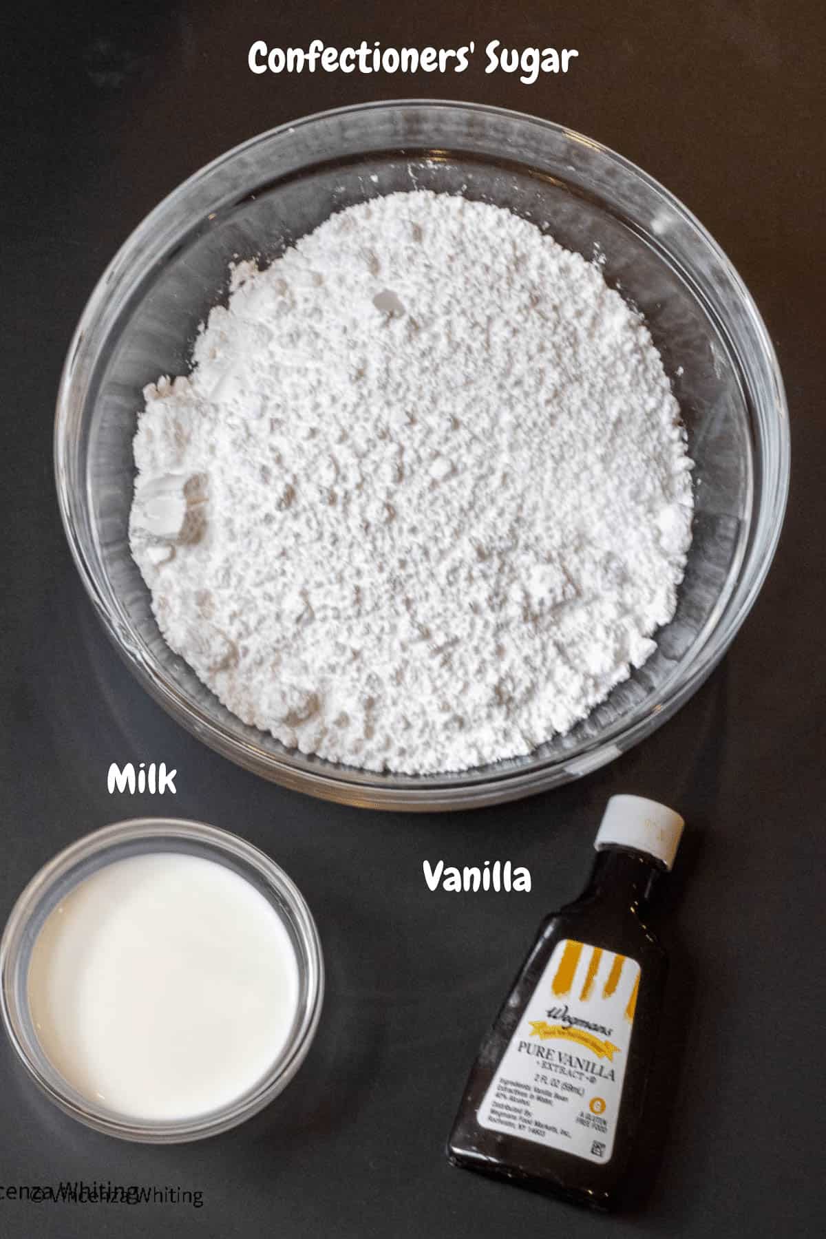 A bowl of confectioner's sugar, a bowl of milk and a small bottle of vanilla extract on a black background.