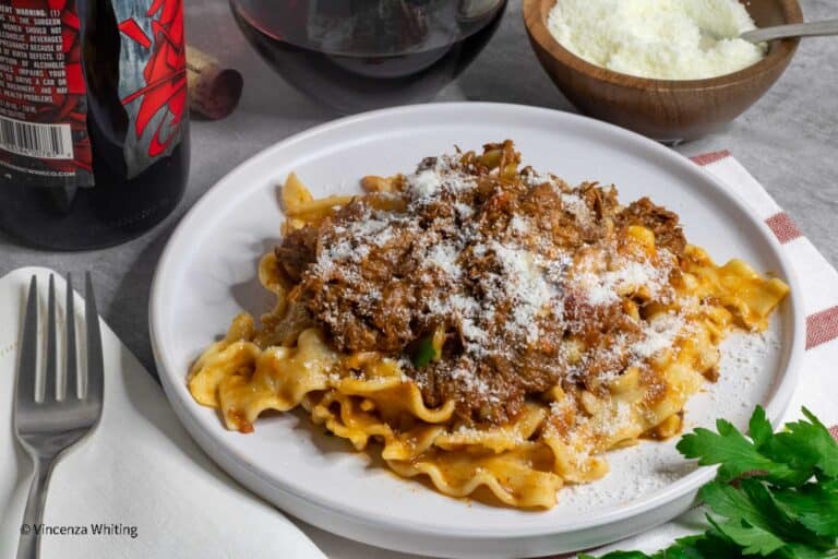 A plate of pasta topped with a hearty meat sauce and sprinkled with grated cheese. A fork and napkin are placed on the left, with a bowl of cheese and a glass of red wine in the background. Fresh parsley decorates the corner.