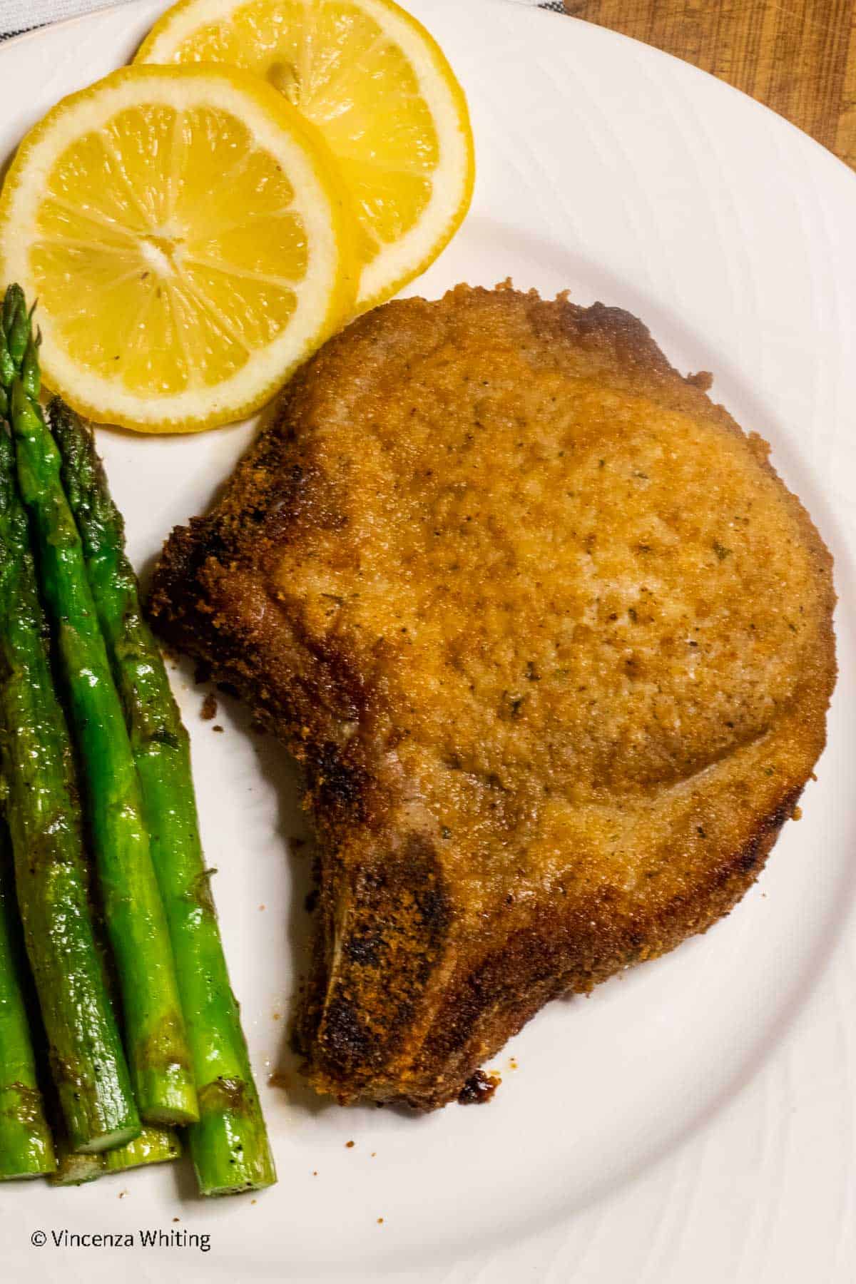 A bone in breaded pork chop with asparagus and lemon slices.