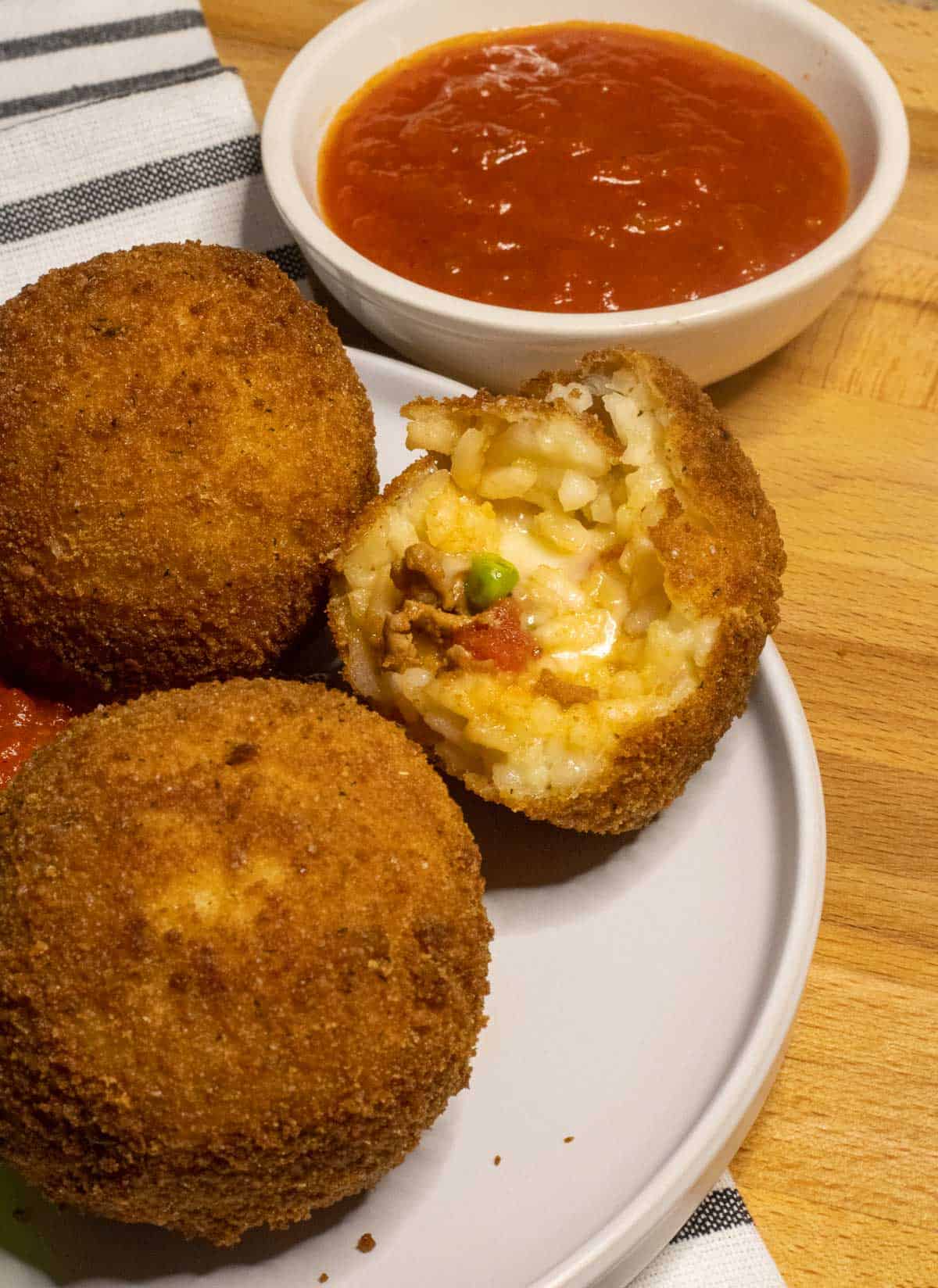 A white plate with breaded and fired rice balls and one is cut in half displaying a meaty sauce and cheese filling.
