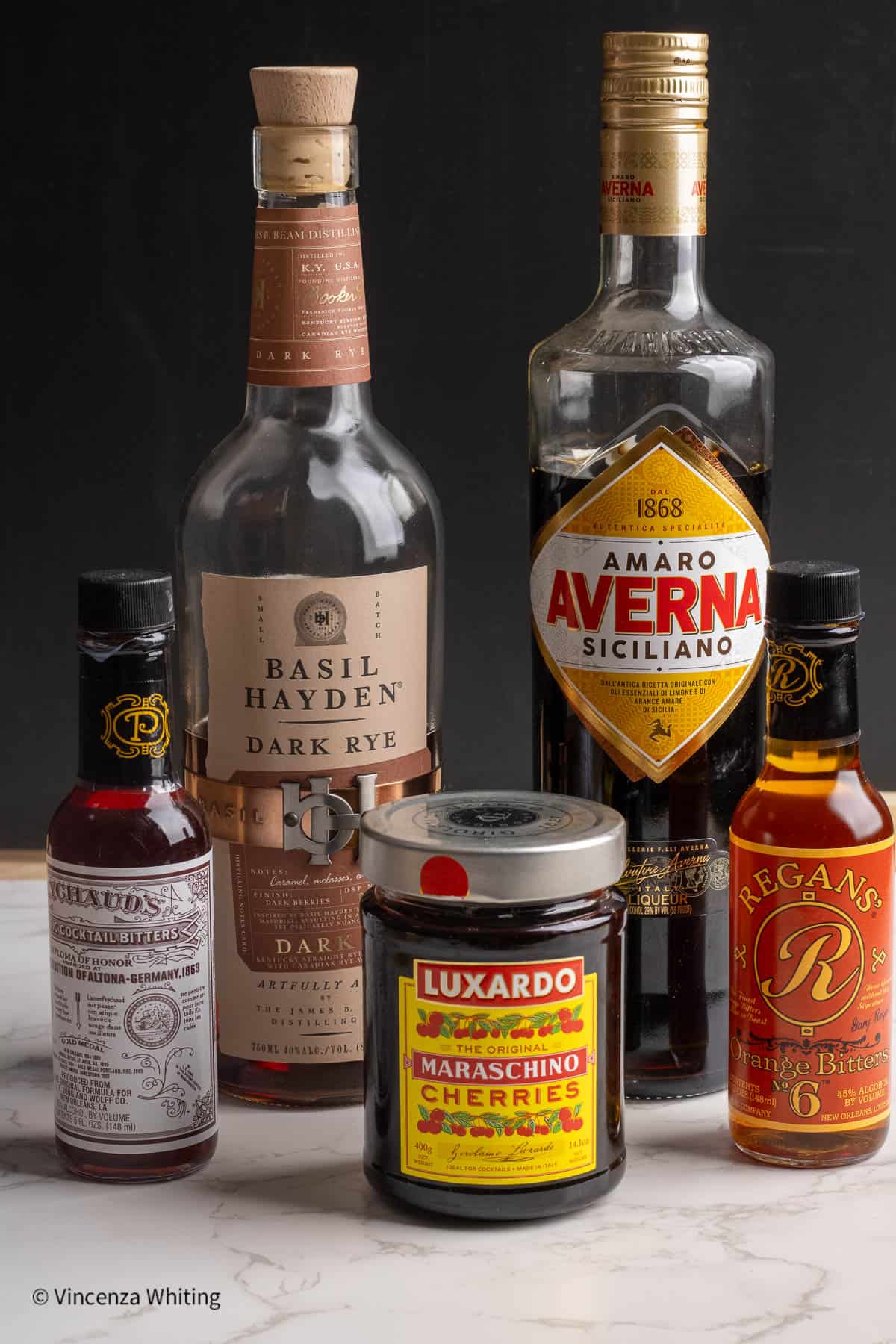 A bottle of Bourbon, a bottle of Amaro, a jar of Luxardo cherries and two bottles of cocktail bitters.