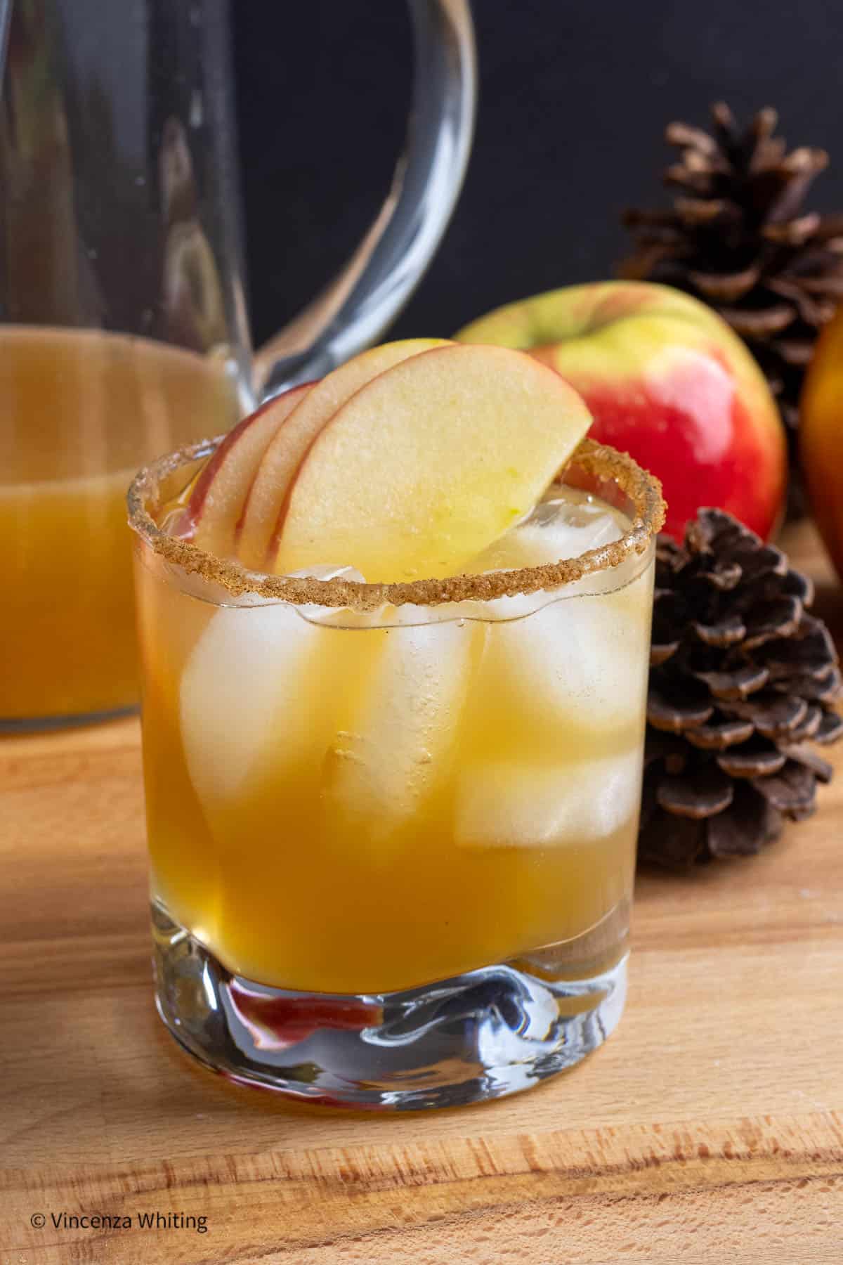 An apple cider cocktail garnished with apple slices and cinnamon sugar rim with pine cones, apples and a pitcher of the drink.