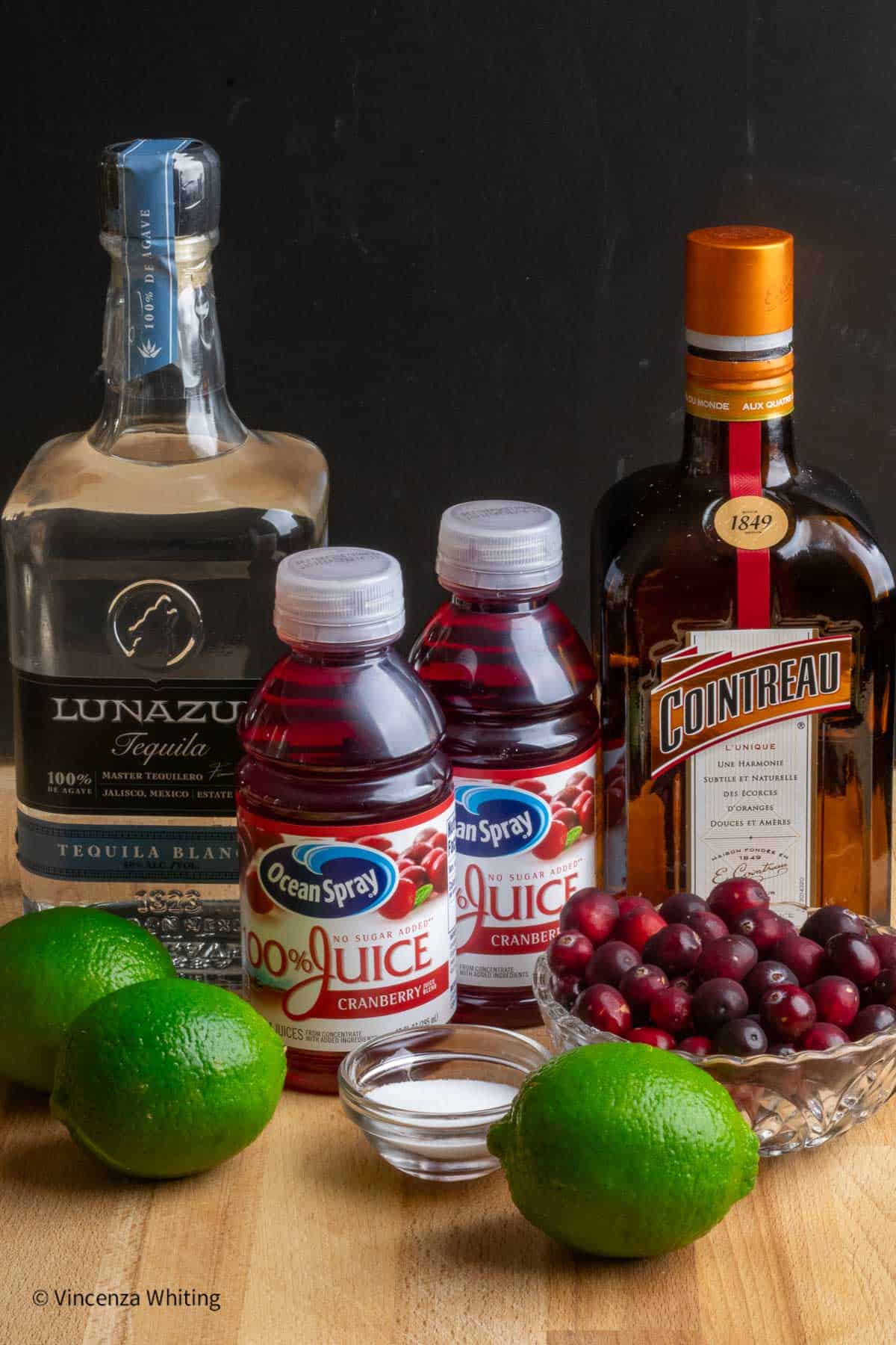 A bottle of tequila, cointreau, limes, cranberry juice, a bowl of cranberries and a bowl of salt.