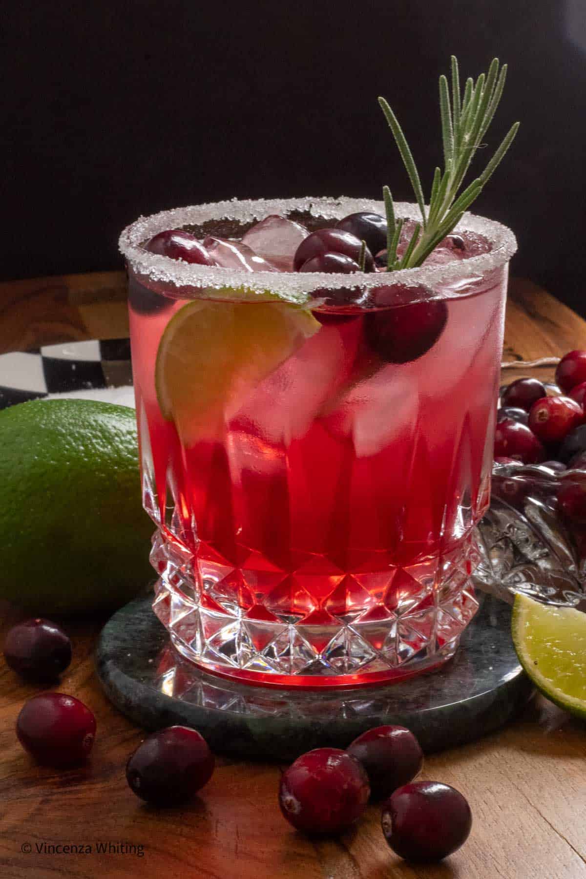 A cranberry margarita garnished with lime, cranberries and a sprig of rosemary surrounded by limes and cranberries.