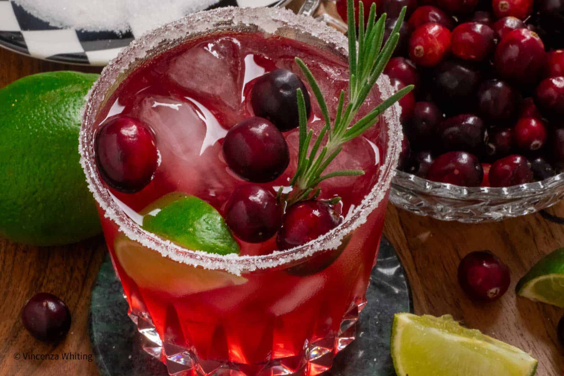 A cranberry beverage with ice garnished with a slice of lime, cranberries and a sprig of rosemary next to a bowl of cranberries and limes.