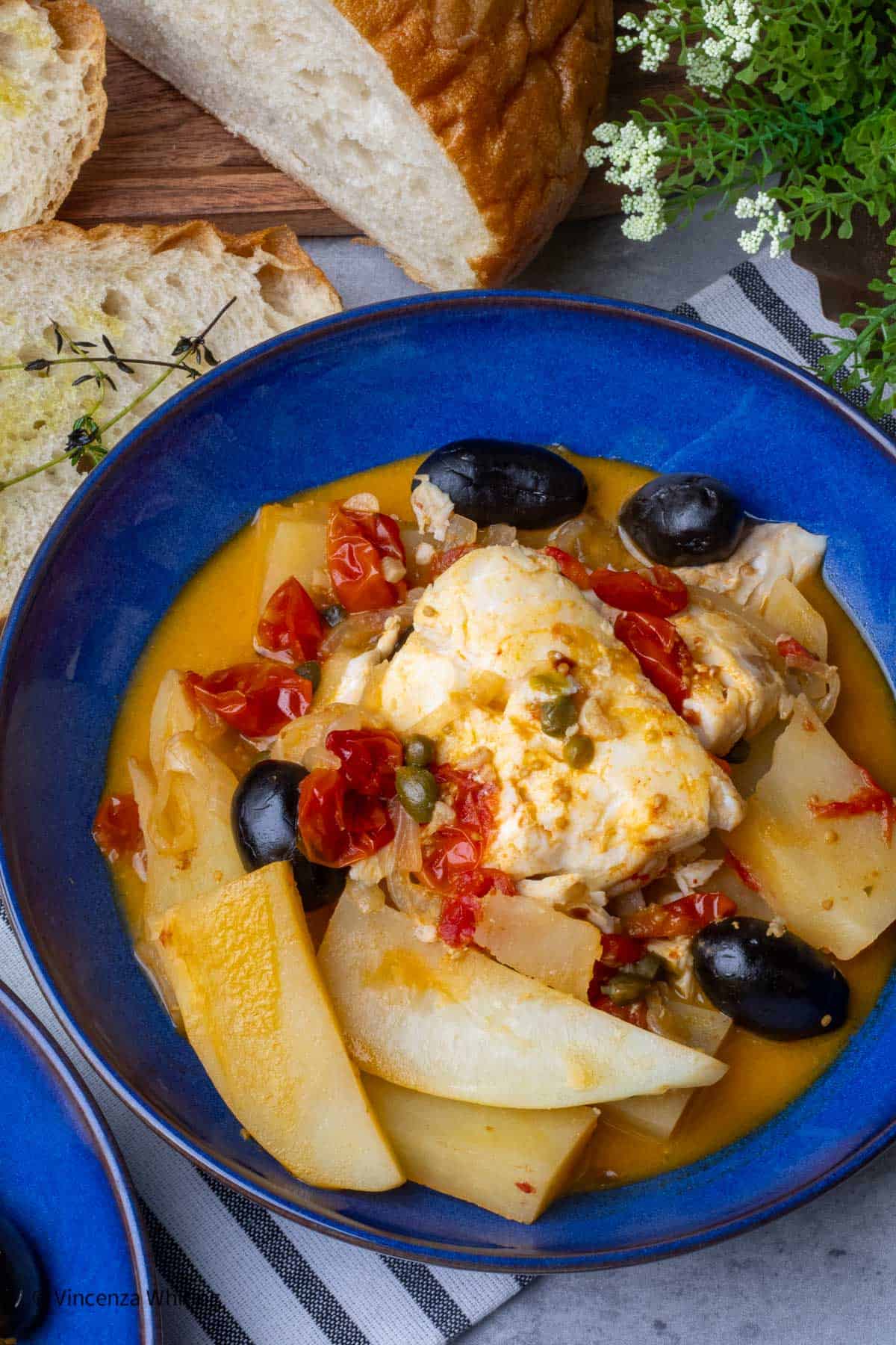A blue bowl of tender white fish in a stew of potatoes, cheery tomatoes and black olives on a table with sliced bread.