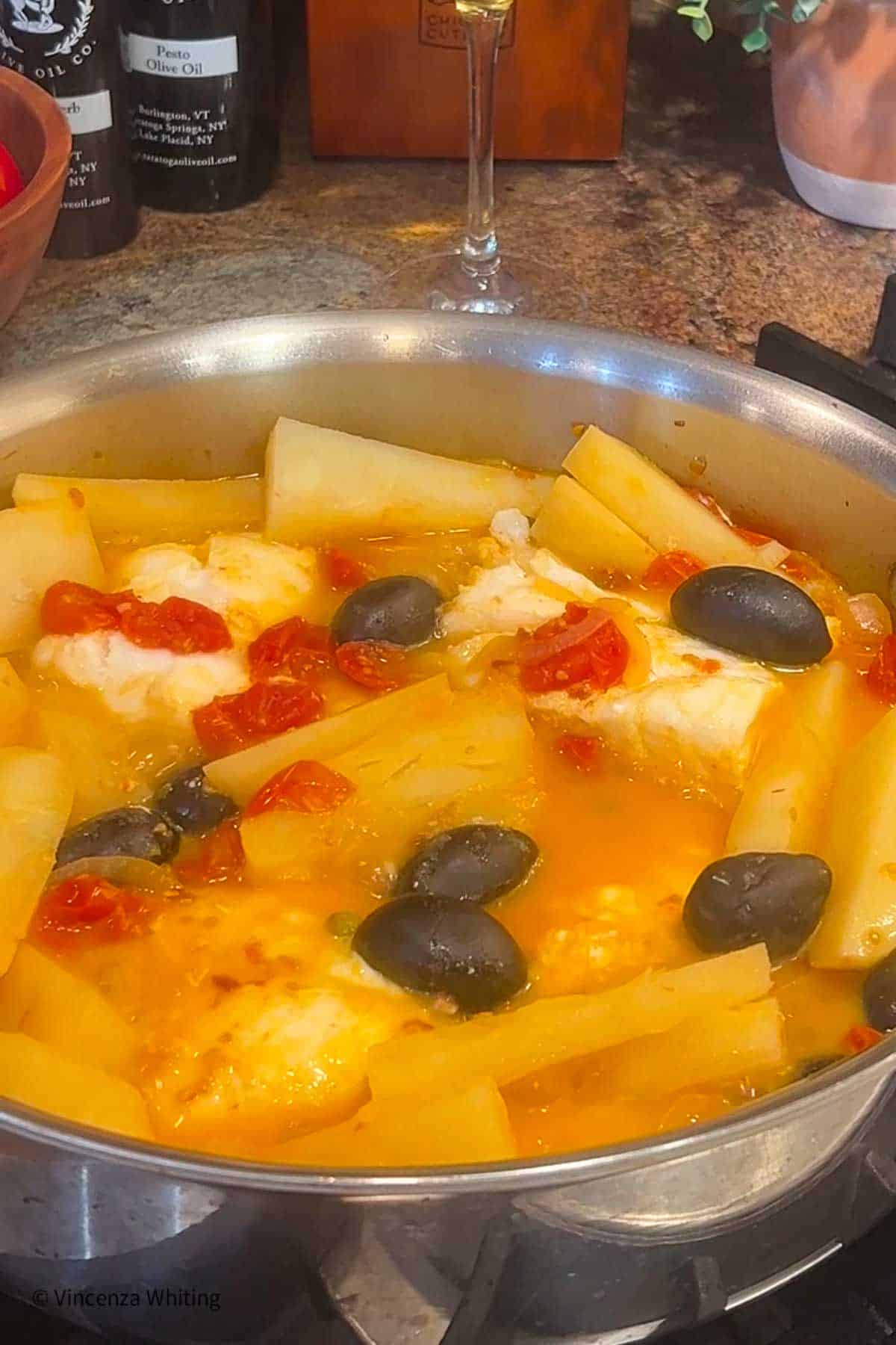 Fish nestled in a sauté pan with potatoes, tomatoes, olives and a broth.