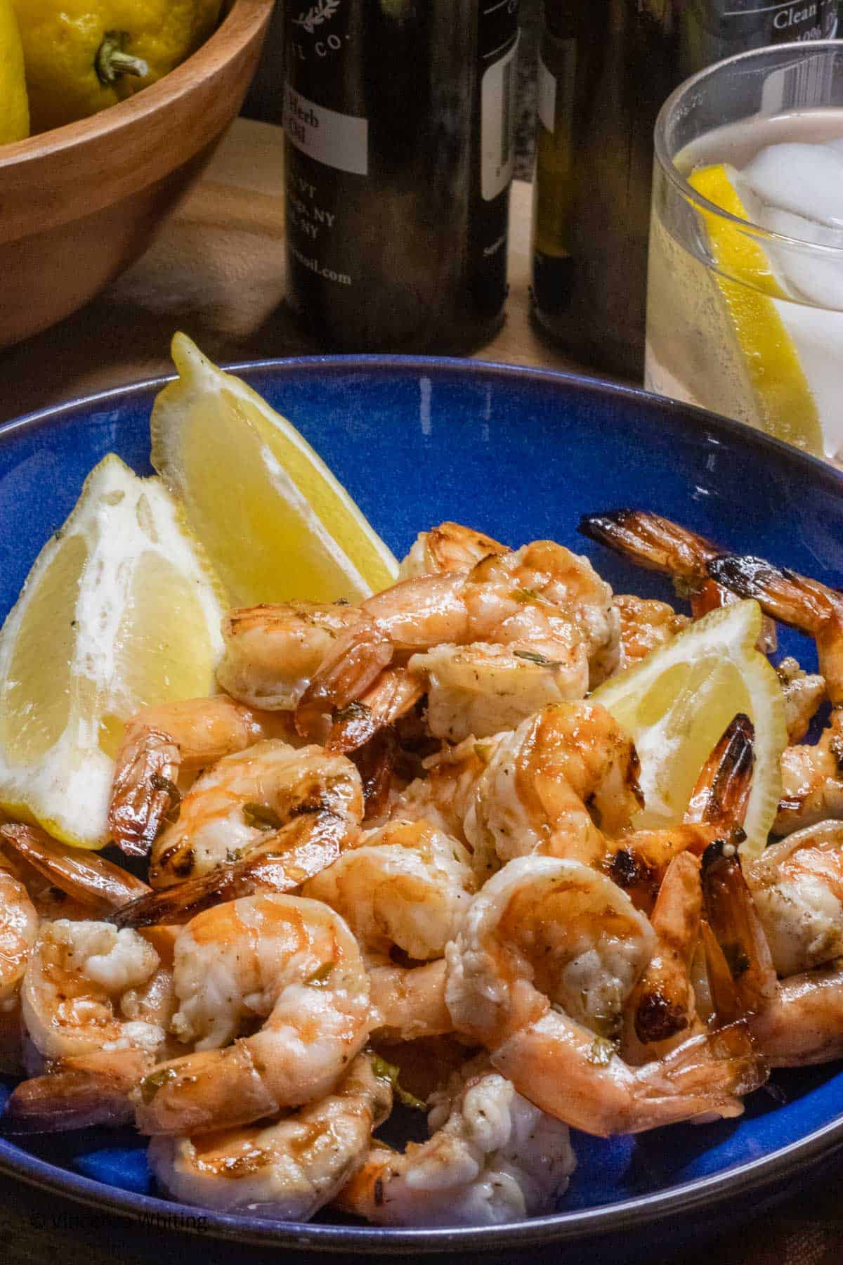 a blue plate with grilled shrimp and lemon wedges in front of a glass and some bottles.
