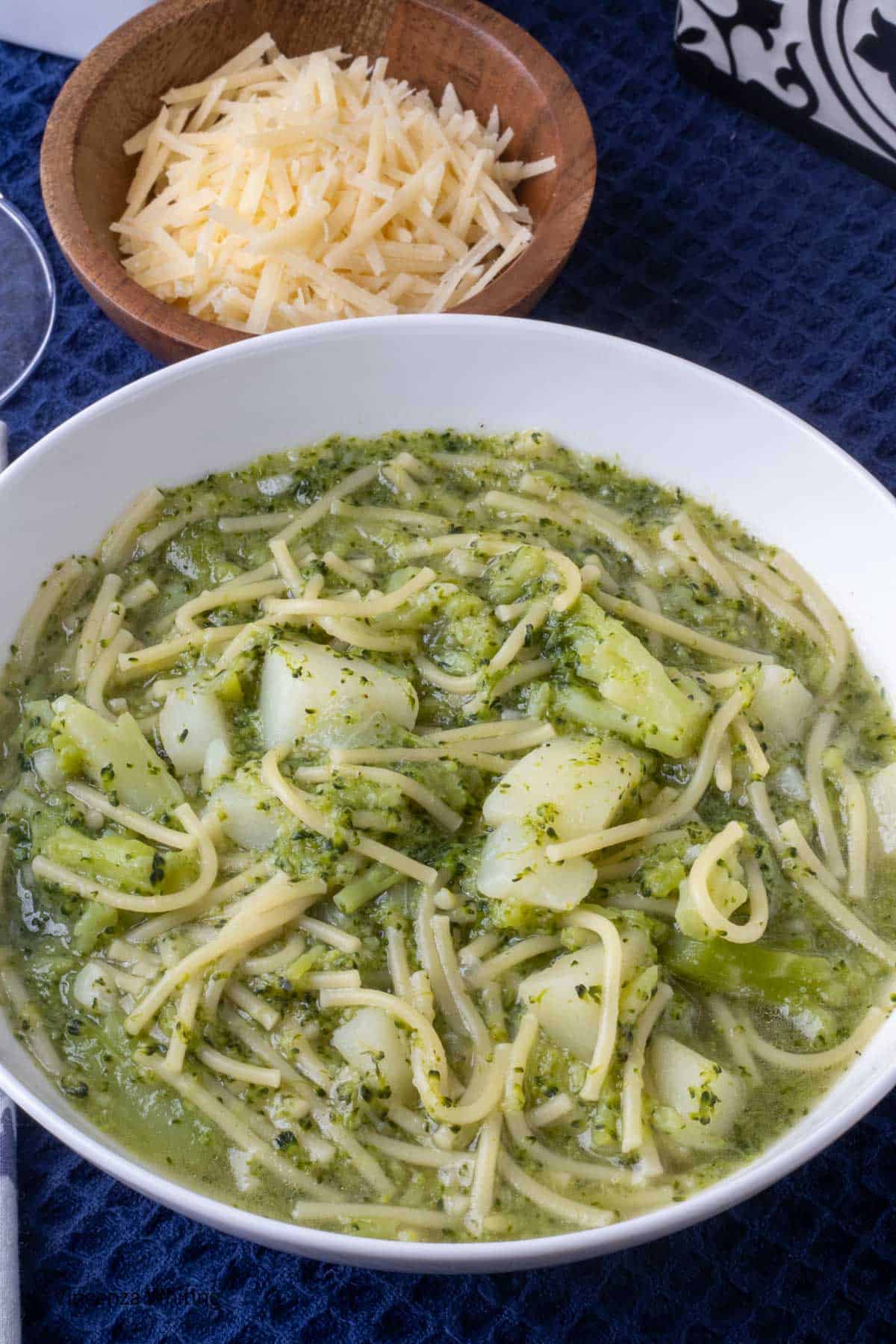 A bowl of broccoli and pasta soup next to a bowl of grated cheese.