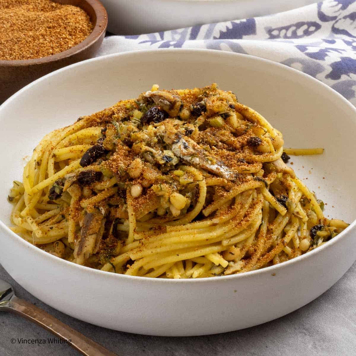 A white bowl of pasta with sardine sauce, topped with toasted breadcrumbs.