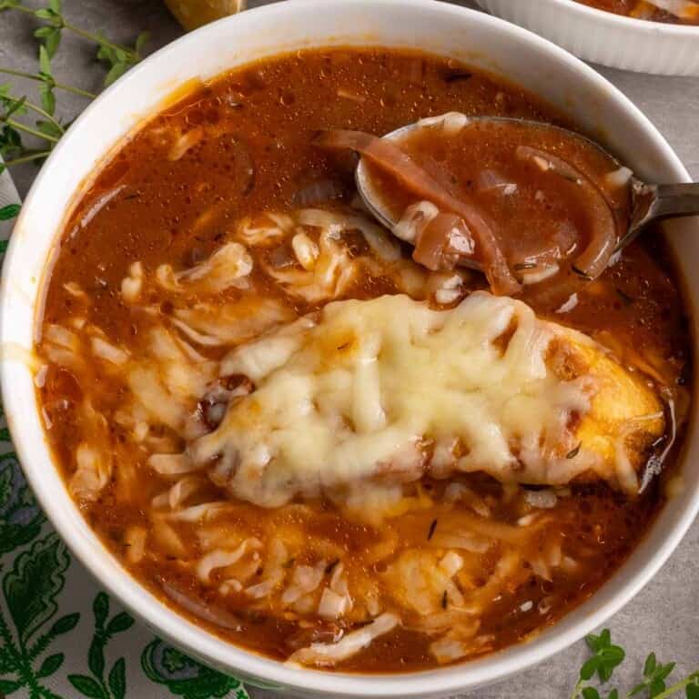 A white bowl filled iwth Italian Onion soup topped with a piece of toasted baguette and melted cheese.