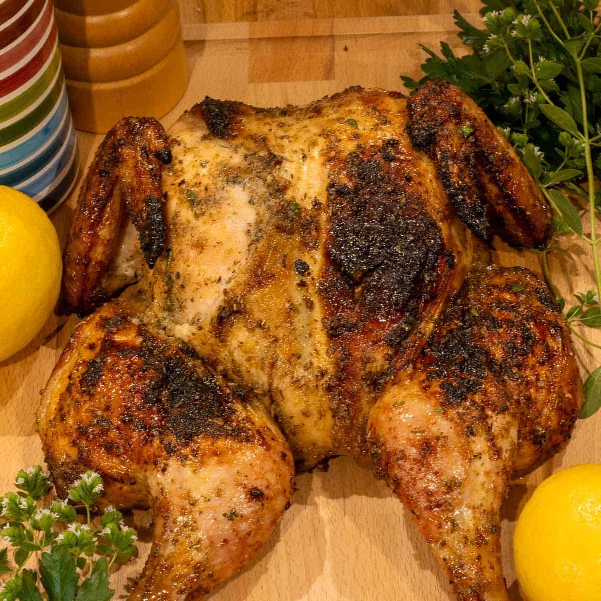 A whole grilled spatchcocked chicken on a cutting board with fresh oregano and parsley.