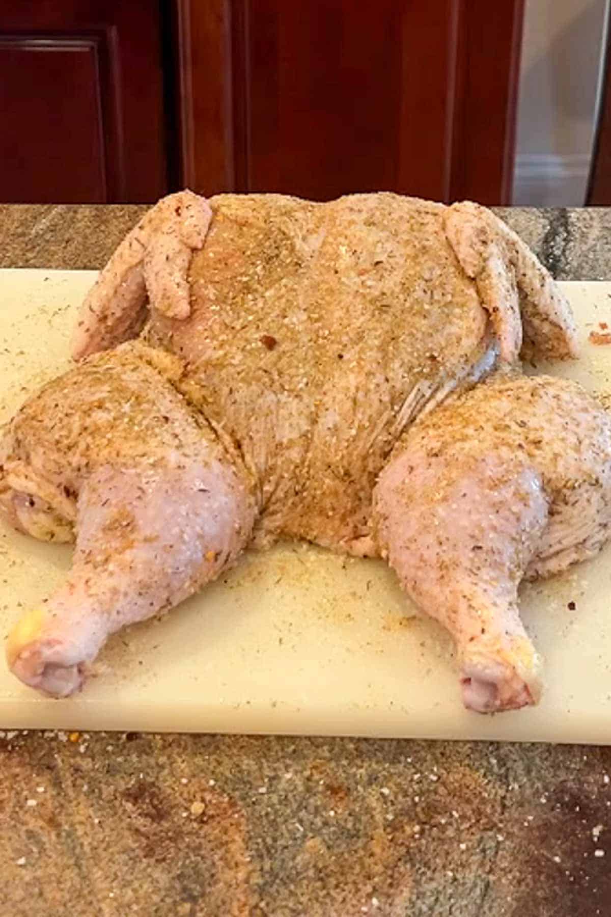 A spatchcocked chicken covered in seasoning on a kwhite cutting board.