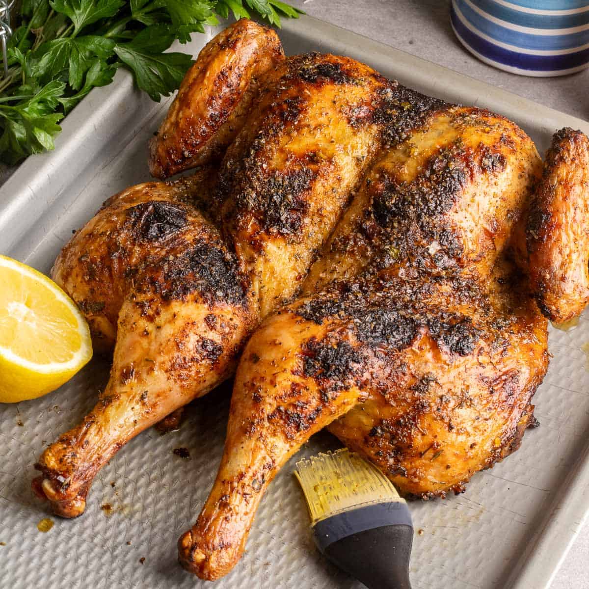 A spatchcocked chicken grilled, on a metal pan with lemons and a basting brush.