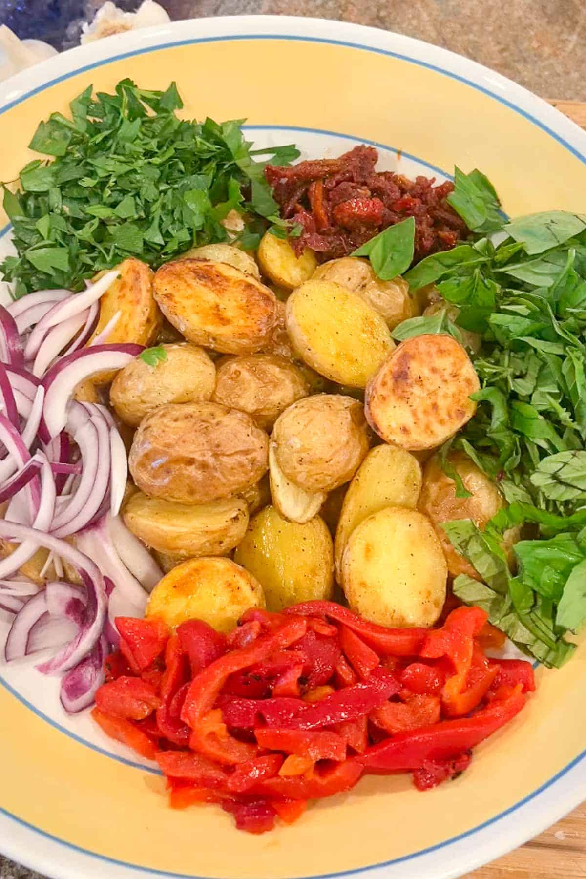 A yellow rimmed bowl filled with roasted baby potato halves, sliced red onion, roasted red pepper strips, chopped parsley and basil and sundried tomato strips.