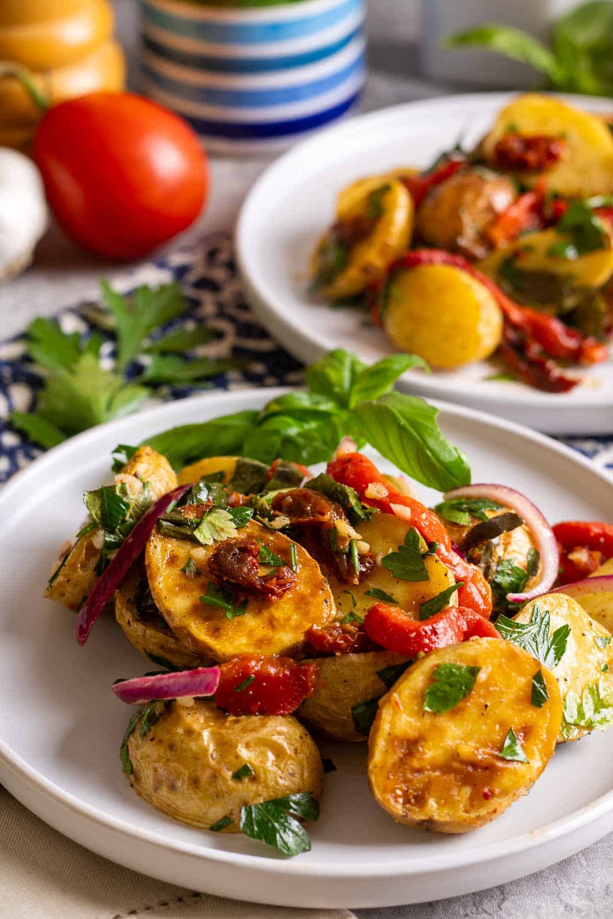 Two white plates of roasted potato salad on a table with parley garlic and a tomato.