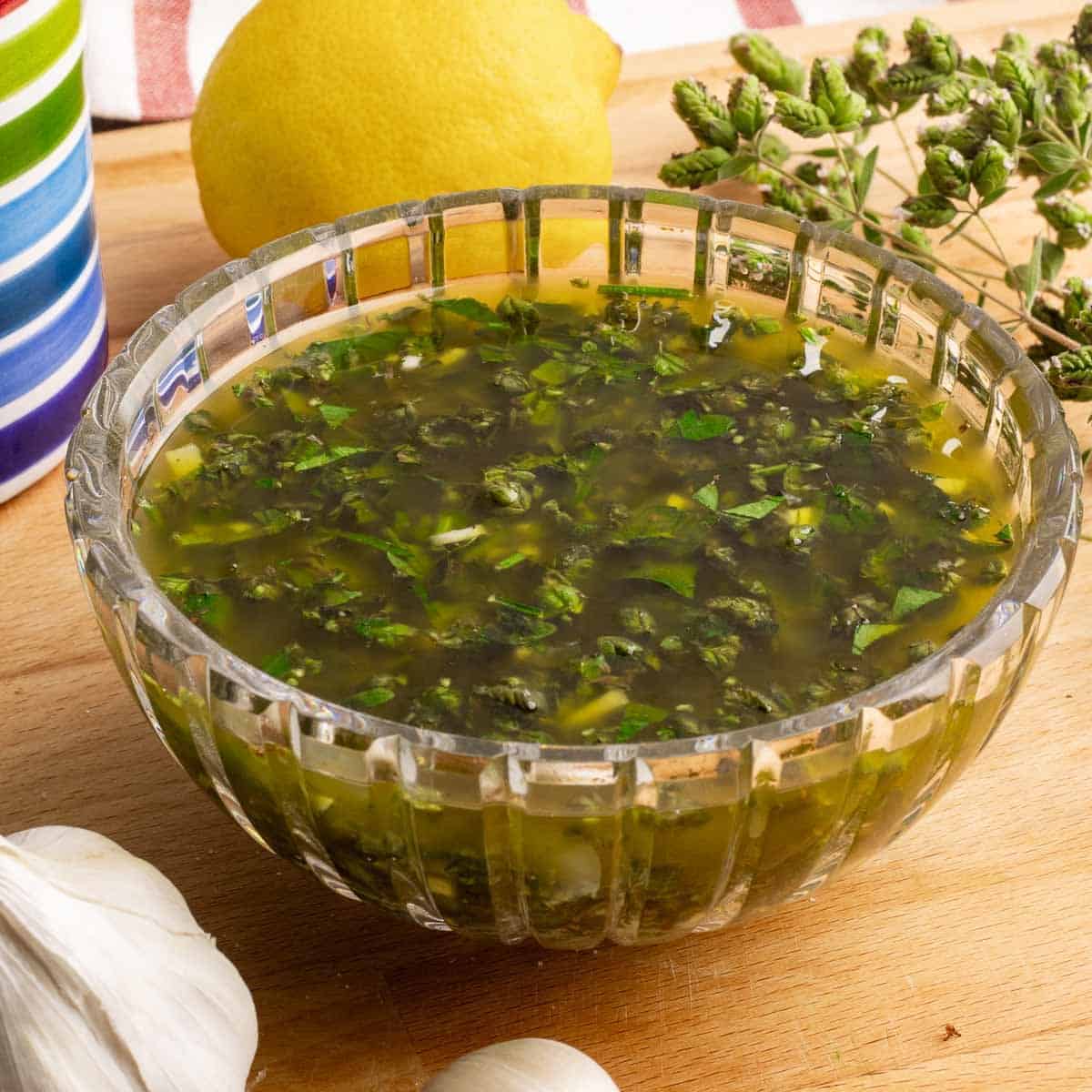 Sicilian Salmoriglio Sauce in a crystal bowl next to lemons garlic and fresh oregano.