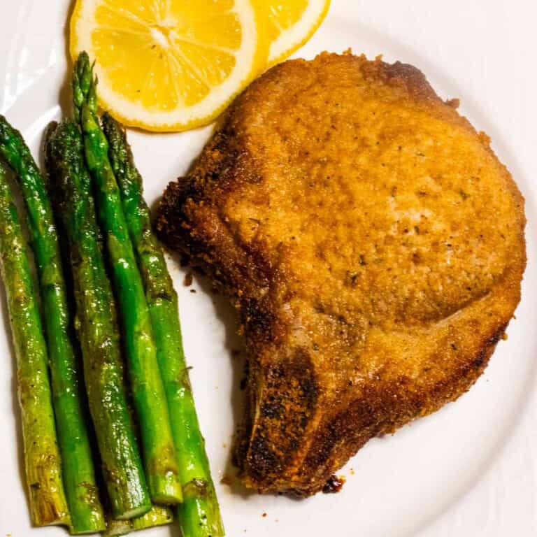 A gloden brown breaded pork chop on a plate with asparagus spears and lemon slices.