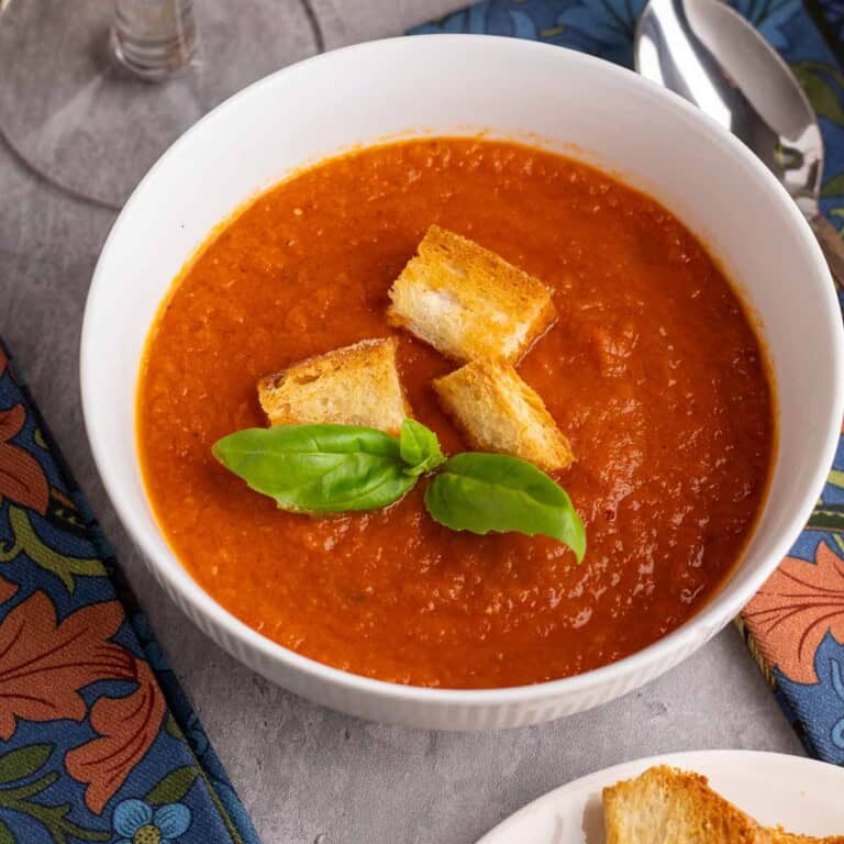 A bowl of roasted tomato and garlic soup with croutons and basil leaves.