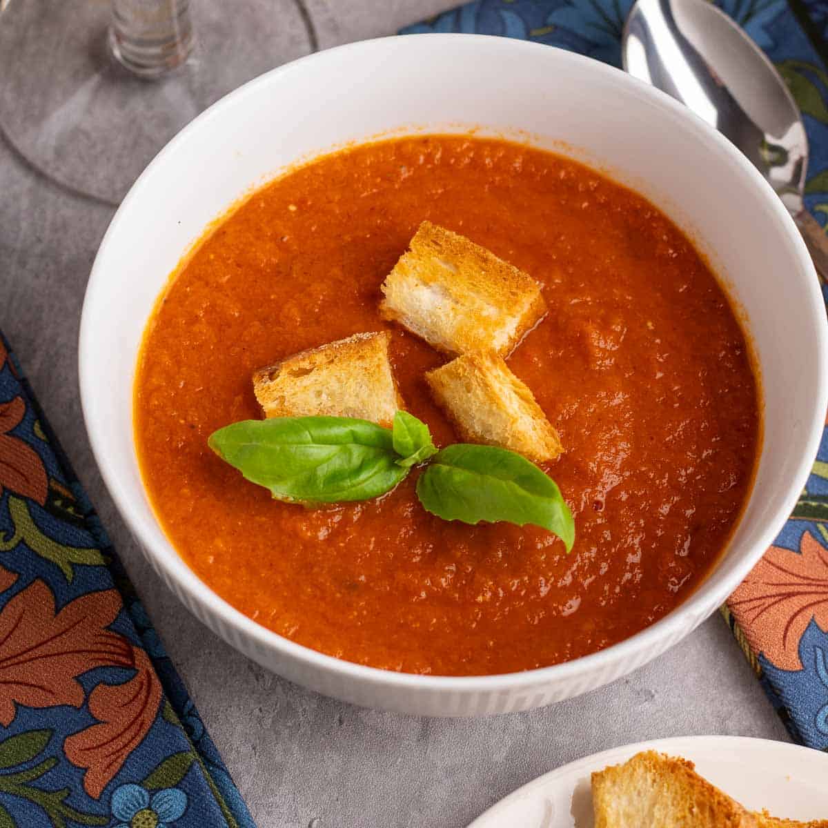 A bowl of roasted tomato and garlic soup with croutons and basil leaves.