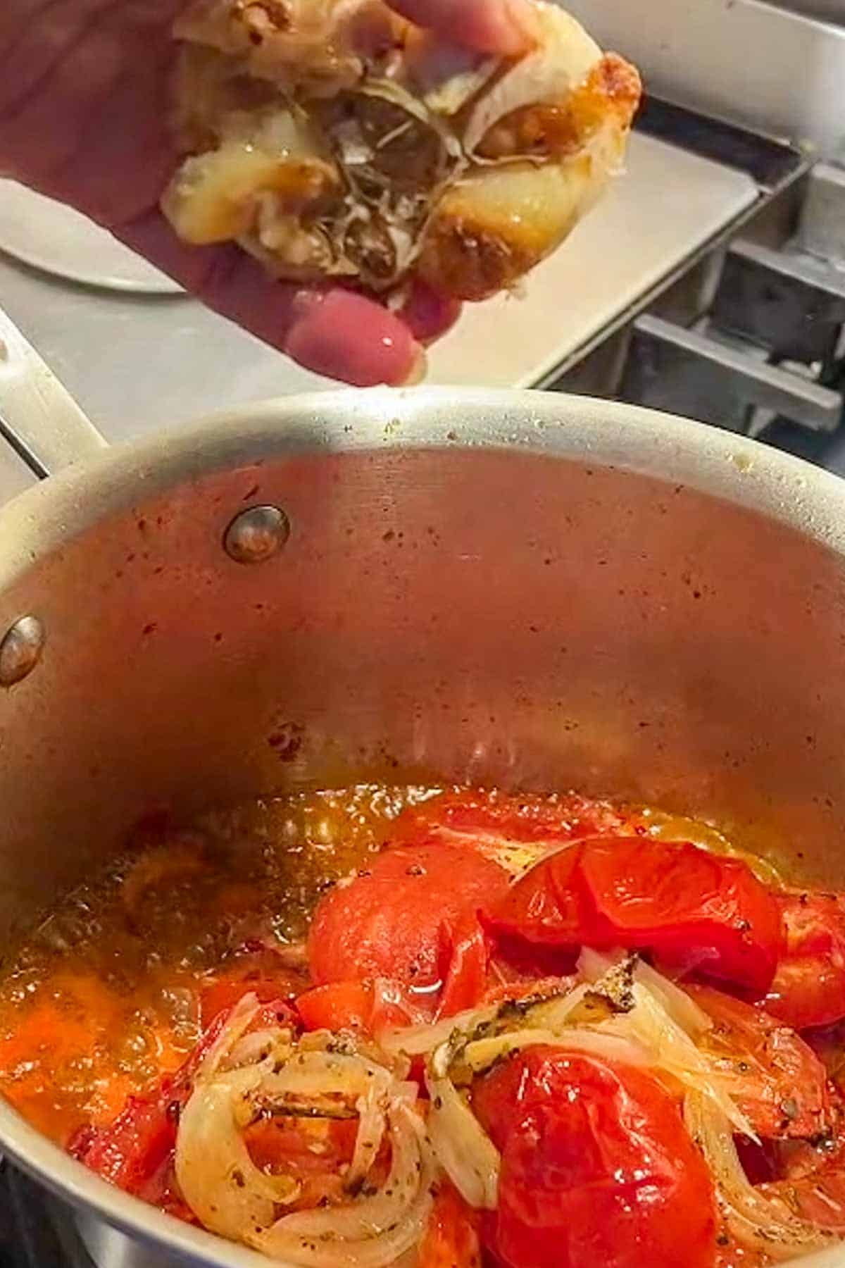 A head of roasted garlic is being squeezed into a pan of tomatoes and onions.