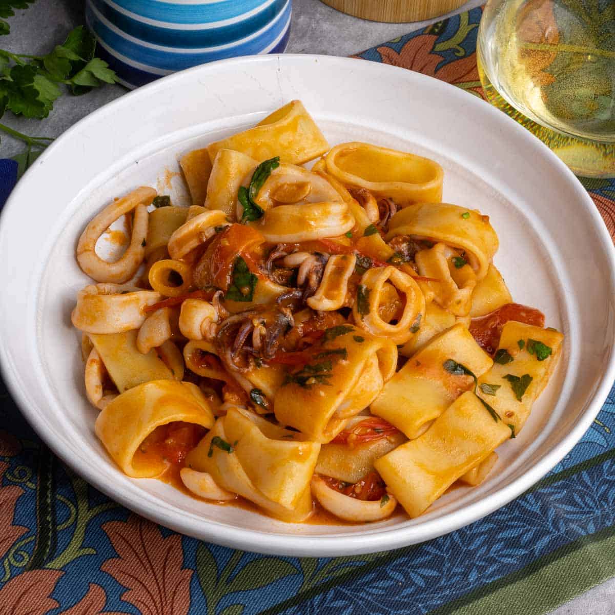 Calamarata pasta with tomato calamari sauce in a white bowl sitting on a colorful towel.