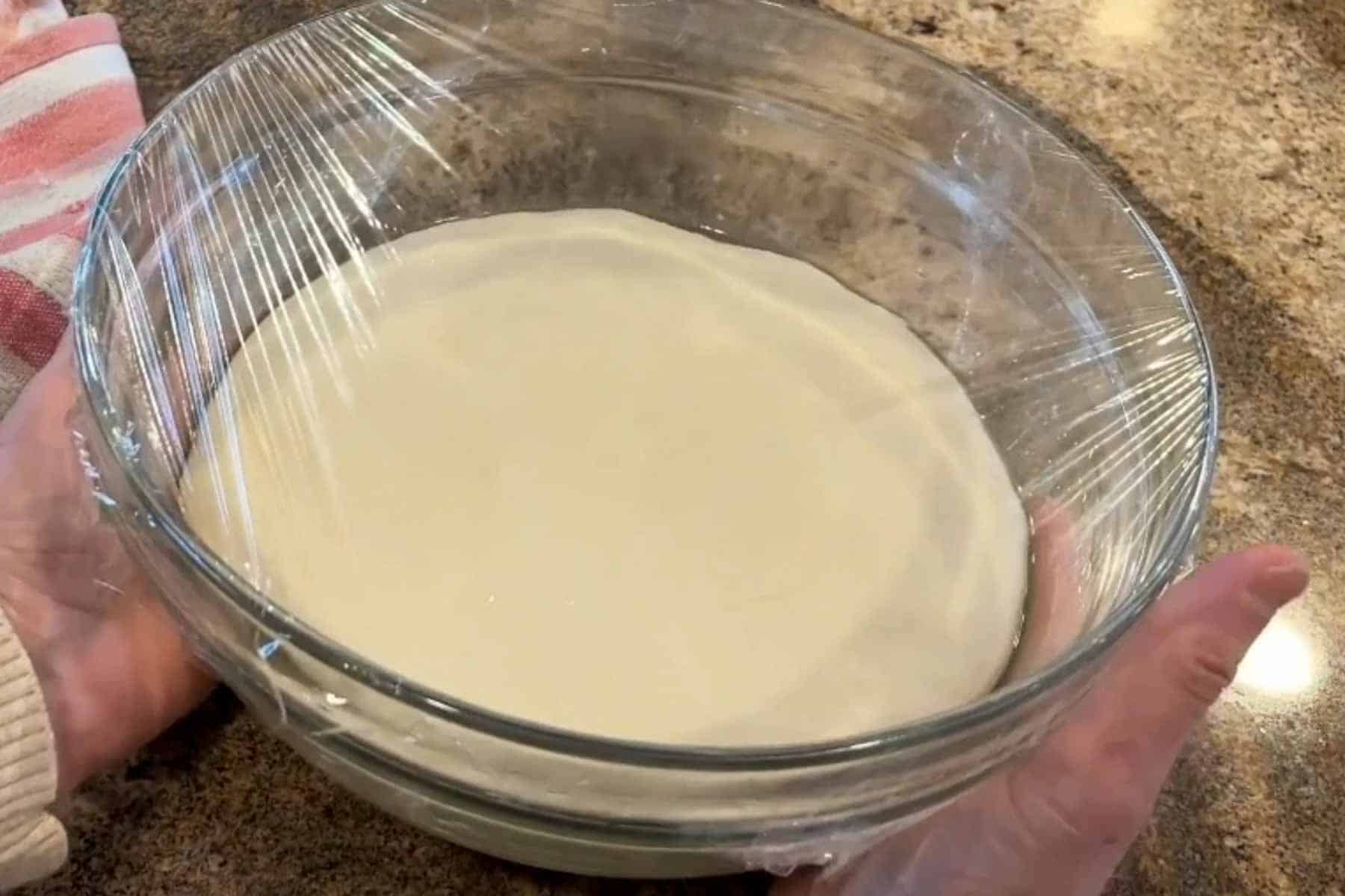 Pinsa Romana dough in a glass bowl covereed with plastic wrap.