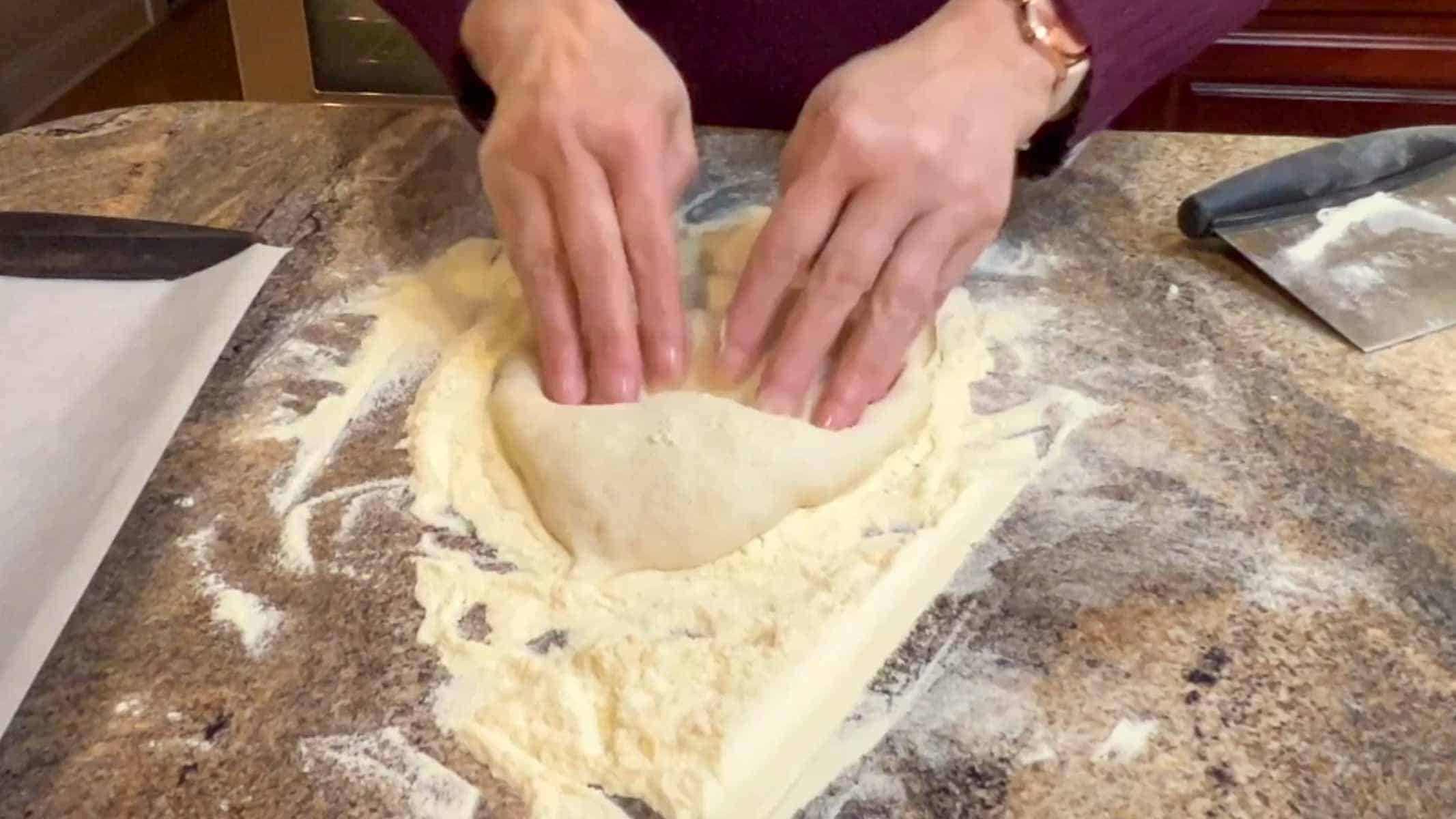 Two hands pushing fingers into dough on a heavily floured counter. 