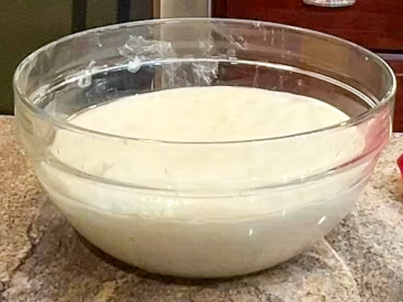 Pinsa Romana dough doubled in size in a glass bowl covered with clear pastic wrap.